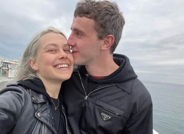 Phoebe Bridgers and Paul Mescal on a pier by the sea, Phoebe smiling as Paul kisses her forehead