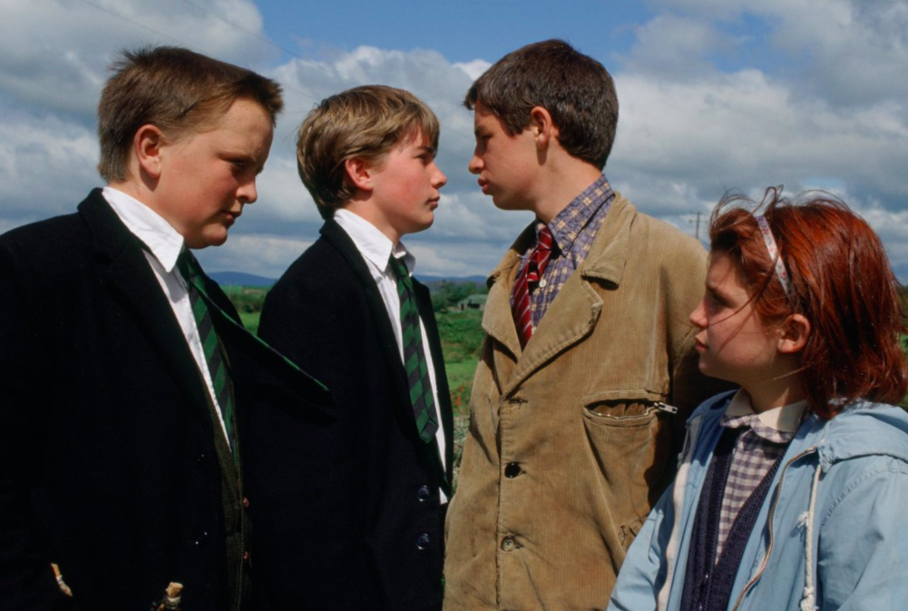 Still from War of the Buttons. Two boys in school uniforms face a taller boy in a brown jacket accompanied by a little girl with red hair.