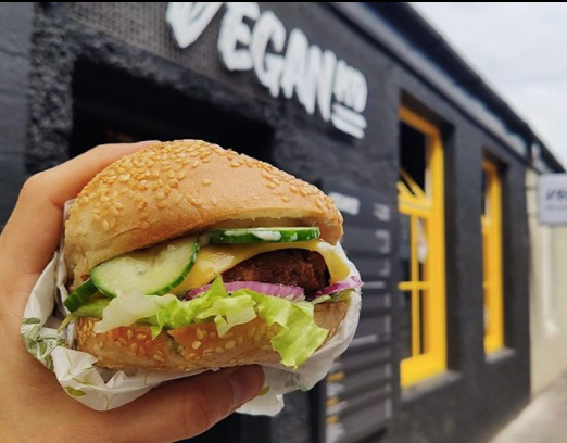 close up of a burger with pickles, cheese and lettuce held up outside a black painted building with yellow windows