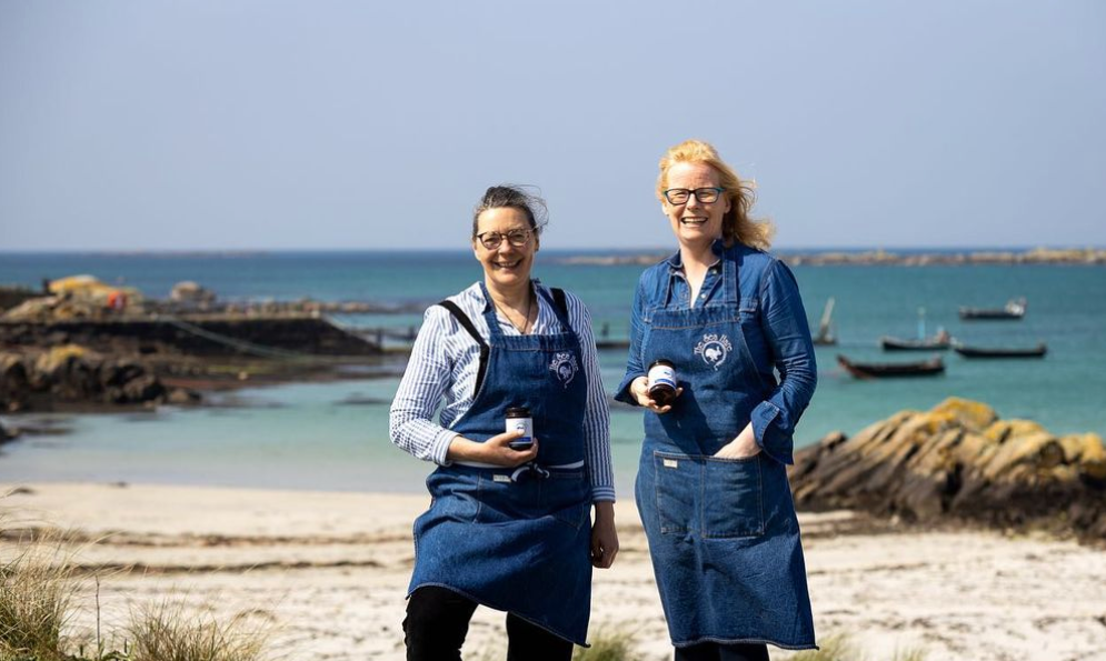 the two owners of the Sea Hare with aprons and coffee cups smiling with a beach in the background