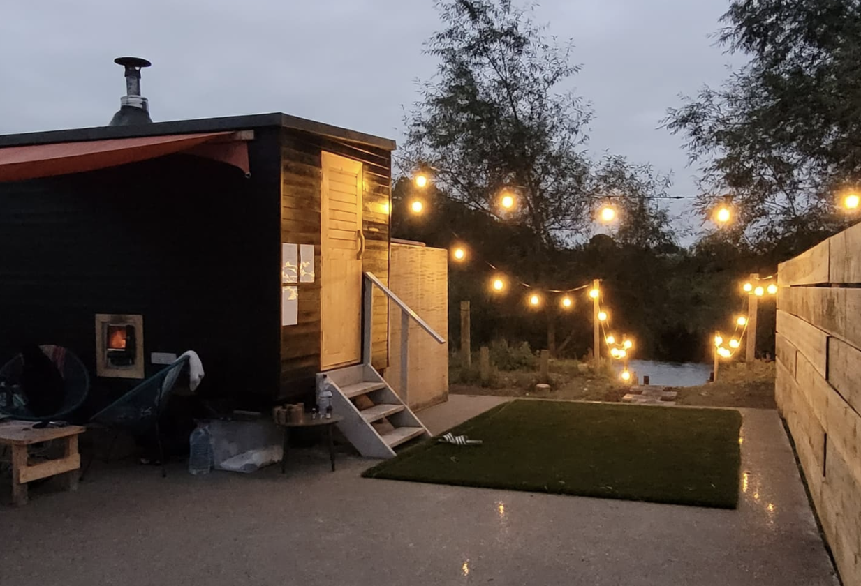 Hot box outdoor spa with sauna to the left, walkway down to a river and fairy lights hanging above