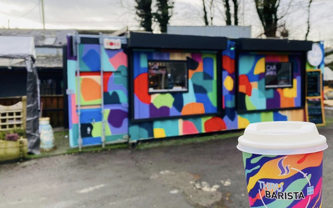 converted shipping container painted with multicoloured shapes, a thru barista cup with similar design is held in the forgeground