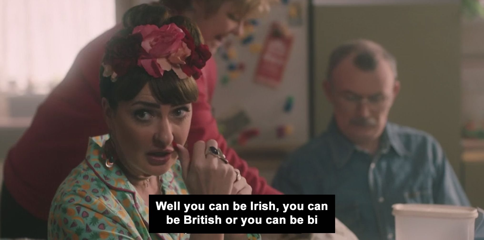 Aunt Sarah from Derry Girls wearing colourful pyjamas and flowers in her hair. Subtitles read - "Well you can be Irish, you can be British or you can be bi"