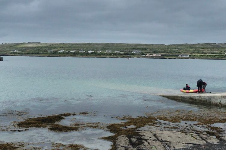 Inis Mor pier with two people pushing a kayak into the water, more green hills and countryside can be seen across the sea in the background