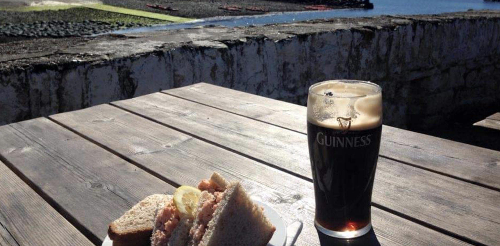 a pint of guinness and a crab sandwich on a picnic bench beside the sea