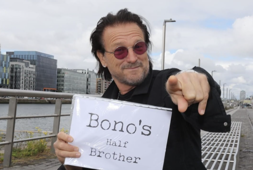 Bono lookalike sitting on a bench by a river holding a sign that reads "Bono's half brother"